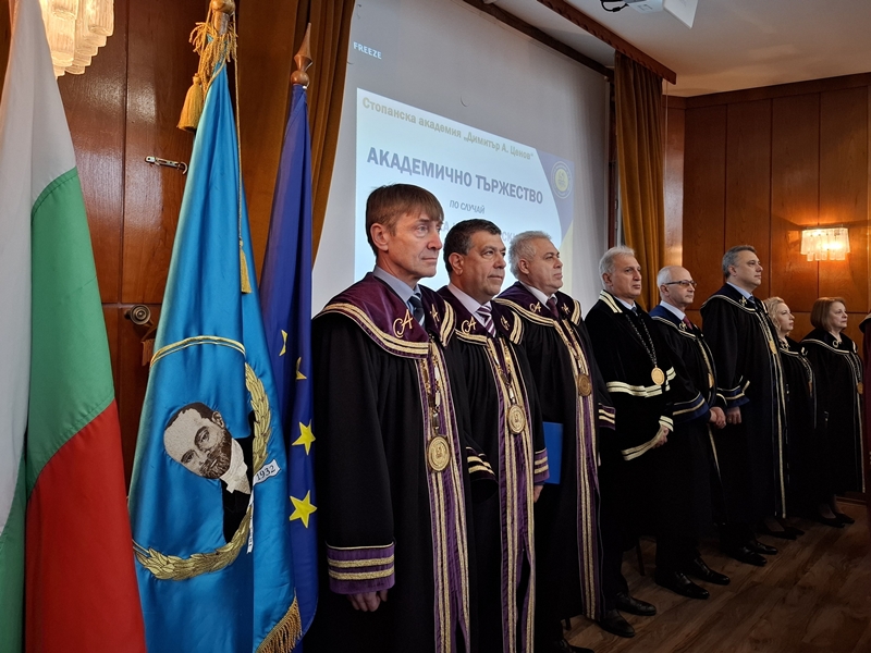 Академичното ръководство облечени в академични роби позират пред стена с надпис отбелязвайки Студентския празник в Стопанска академия – Свищов