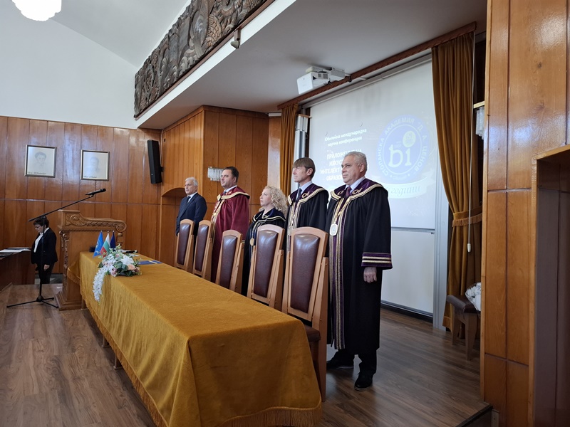 Официална церемония в дървен панелен салон с хора в академични тоги и дълга маса покрита със зелена покривка. Виждат се поздравителни плакети и цветя