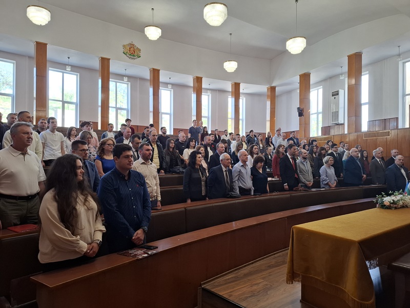 Голяма група хора седящи в просторна зала с високи тавани празнуват 60-годишнината на катедра „Бизнес информатика“. На масата в предния план има цветя и украса