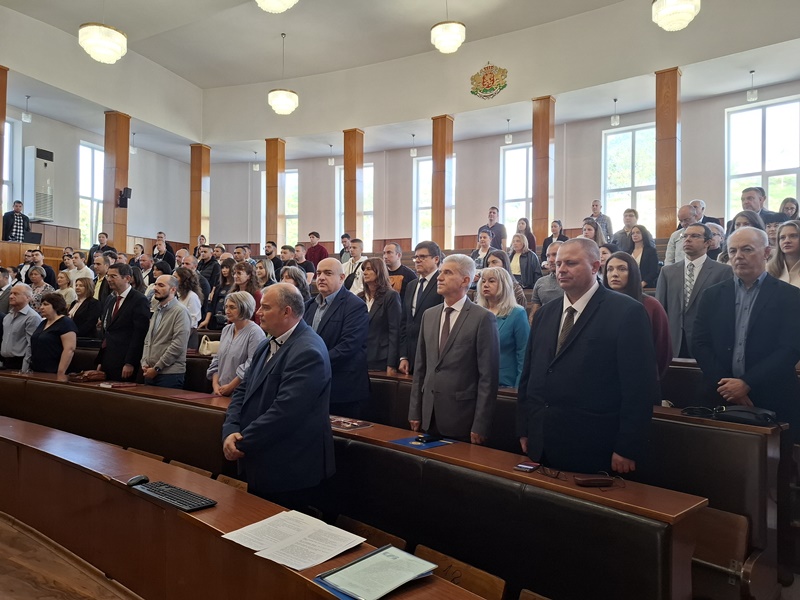Голяма група хора в официално облекло присъстват на честване в светла и просторна зала. Виждат се дървени пейки и осветление